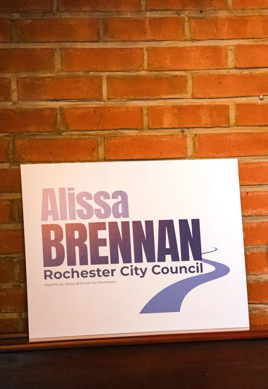 An Alissa Brennan for Rochester City Council lawn sign leaning against a brick wall at the campaign launch